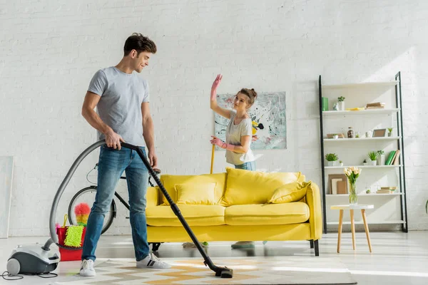 Glücklicher Mann mit Staubsauger in der Nähe einer winkenden Frau im Wohnzimmer — Stockfoto