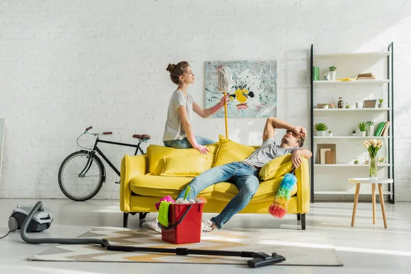 Истощенный мужчина сидит на диване рядом ведро с домашним хозяйством и женщиной — стоковое фото