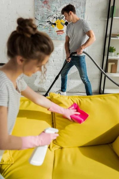 Selective focus of handsome man using vacuum cleaner near girl washing sofa with rag — Stock Photo