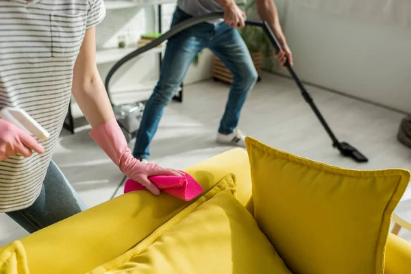 Vista cortada de mulher segurando pano e spray perto de sofá e homem de limpeza sala de estar com aspirador de pó — Fotografia de Stock