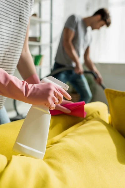 Foco seletivo da mulher segurando pano e spray garrafa perto de sofá e homem na sala de estar — Fotografia de Stock