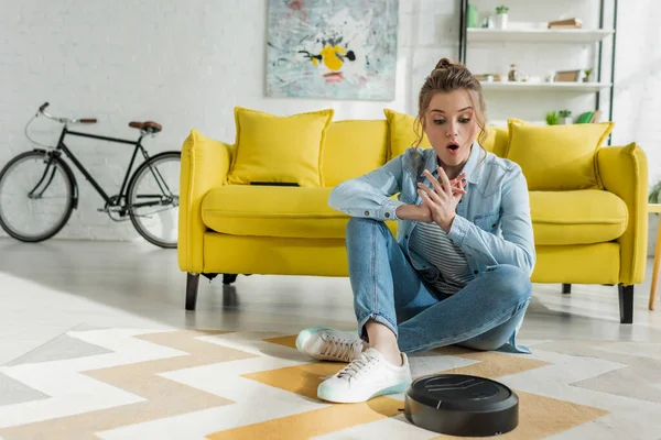 Surprised girl looking at robotic vacuum cleaner on carpet in living room — Stock Photo