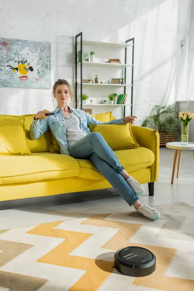 Young woman watching tv while robotic vacuum cleaner washing carpet in living room — Stock Photo