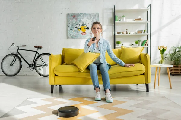 Attraente donna guardando la tv mentre robot aspirapolvere tappeto di lavaggio in soggiorno — Foto stock