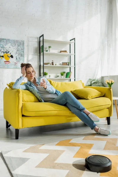 Menina atraente usando smartphone enquanto robótico aspirador de pó tapete de lavagem na sala de estar — Fotografia de Stock