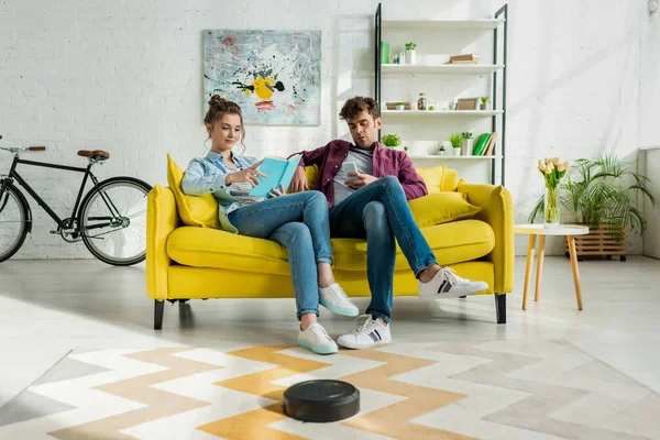 Man using smartphone and woman reading book while robotic vacuum cleaner washing carpet in living room — Stock Photo