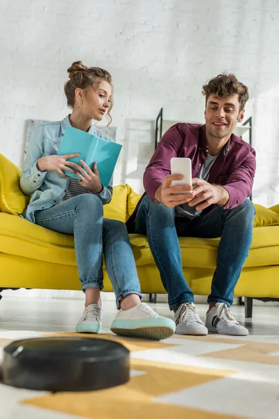 Enfoque selectivo de hombre feliz utilizando el teléfono inteligente y la mujer sosteniendo libro mientras que la aspiradora robótica alfombra de lavado en la sala de estar - foto de stock