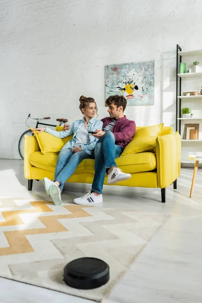 Felice uomo e donna guardando film mentre robot aspirapolvere tappeto di lavaggio in soggiorno — Foto stock