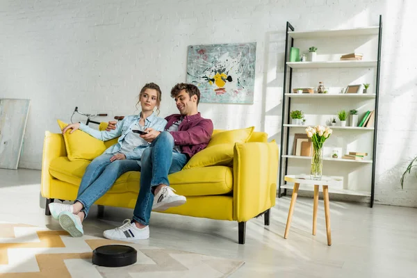 Glücklicher Mann und junge Frau sehen Film, während Staubsaugerroboter Teppich im Wohnzimmer waschen — Stockfoto