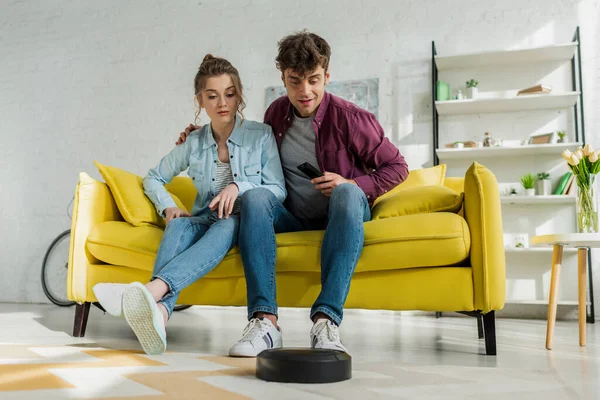 Man and young woman looking at robotic vacuum cleaner washing carpet in living room — Stock Photo