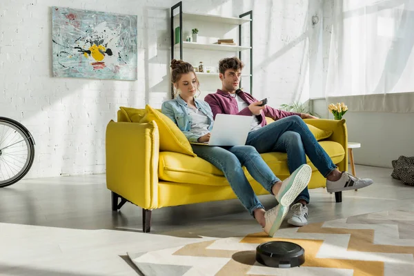 Man watching movie near woman using laptop while robotic vacuum cleaner washing carpet in living room — Stock Photo