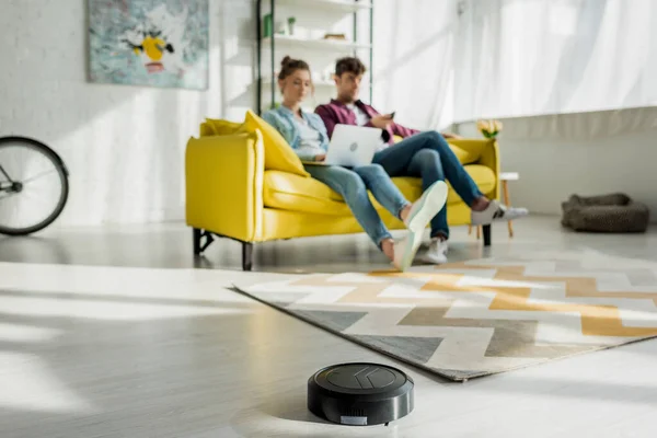 Selektiver Fokus von Staubsaugerroboter-Waschteppich in der Nähe von Mann und Frau, die Laptop im Wohnzimmer benutzen — Stockfoto