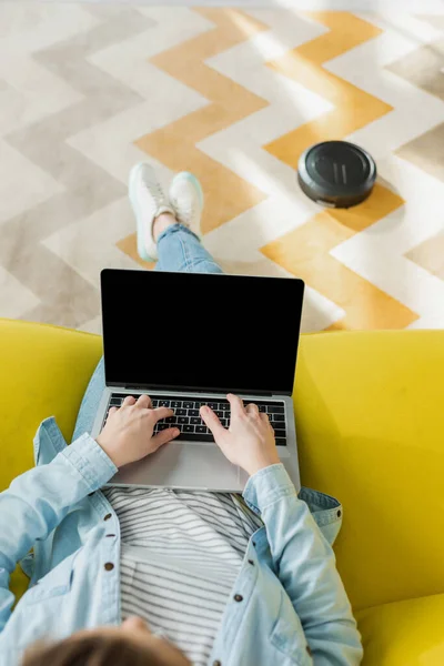 Верхний вид девушки с помощью ноутбука с чистым экраном в то время как роботизированный пылесос стиральный ковер в гостиной — стоковое фото