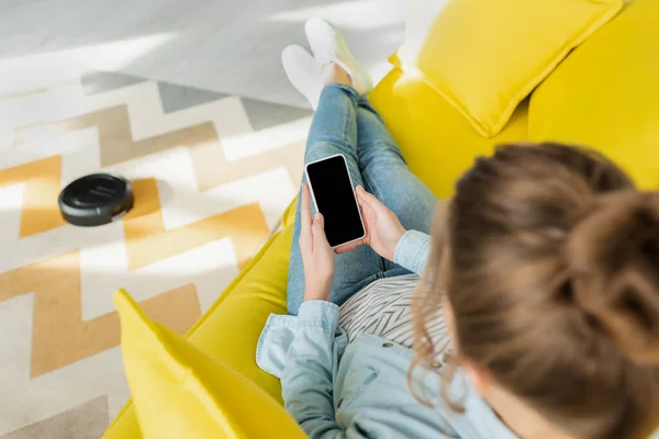 Vista superior de la mujer sosteniendo teléfono inteligente con pantalla en blanco, mientras que la aspiradora robótica alfombra de lavado en la sala de estar - foto de stock