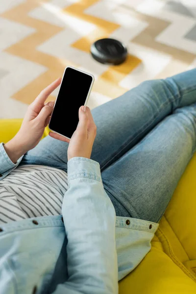 Vista recortada de la mujer sosteniendo teléfono inteligente con pantalla en blanco, mientras que la aspiradora robótica alfombra de lavado en la sala de estar - foto de stock