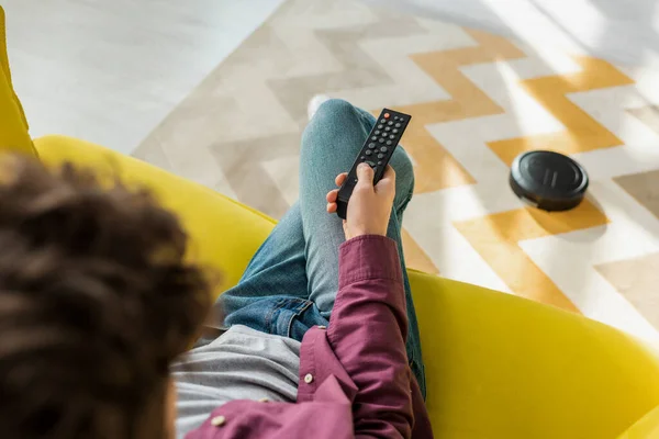 Selective focus of man sitting on sofa and watching movie while robotic vacuum cleaner washing carpet in living room — Stock Photo