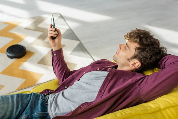 Vista aérea del hombre relajándose en el sofá y viendo películas mientras la aspiradora robótica lava la alfombra en la sala de estar - foto de stock
