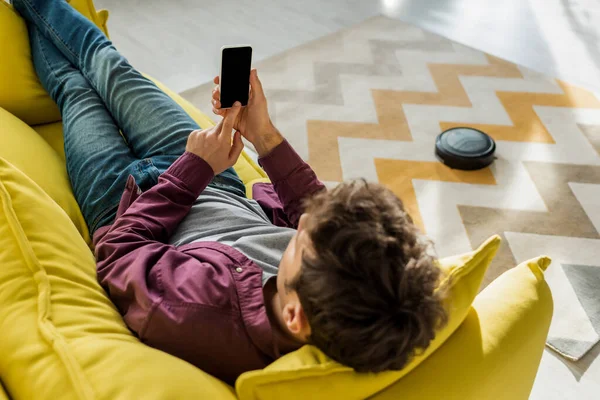 Vista aérea do homem relaxante no sofá e segurando smartphone com tela em branco, enquanto robótico aspirador de pó tapete de lavagem na sala de estar — Fotografia de Stock