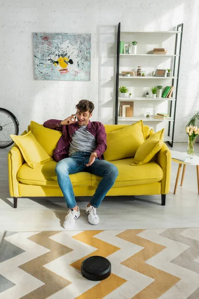 Happy man talking on smartphone while robotic vacuum cleaner washing carpet in living room — Stock Photo