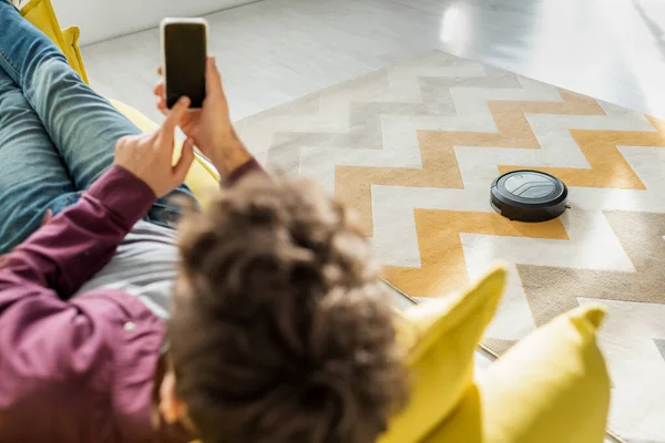 Selektiver Fokus des Mannes, der sich auf dem Sofa entspannt und mit dem Finger auf Smartphone mit leerem Bildschirm zeigt, während Staubsaugerroboter Teppich waschen — Stockfoto