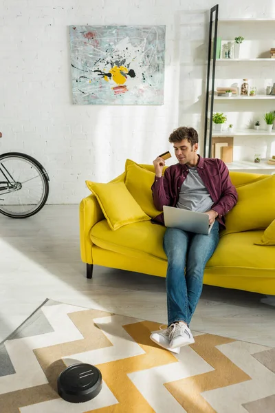 Handsome man online shopping while robotic vacuum cleaner washing carpet in living room — Stock Photo