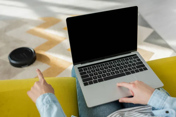 Cropped view of woman pointing with finger at robotic vacuum cleaner near laptop with blank screen — Stock Photo