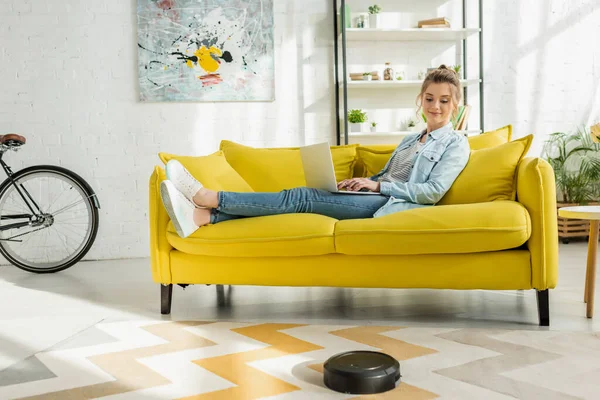 Mujer feliz usando el ordenador portátil, mientras que la aspiradora robótica alfombra de lavado en la sala de estar - foto de stock
