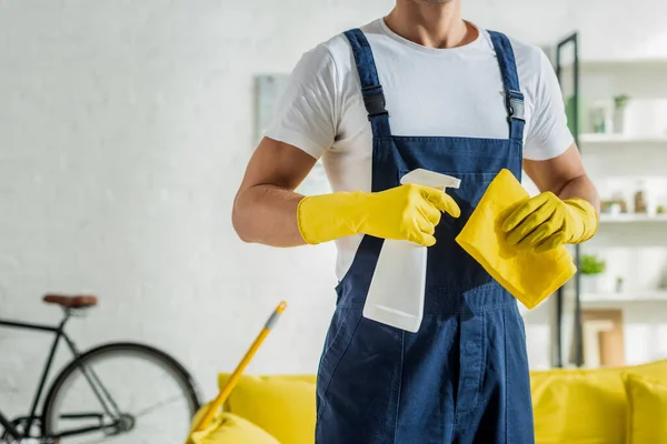 Cropped view of cleaner in overalls holding rag and spray bottle in living room — Stock Photo