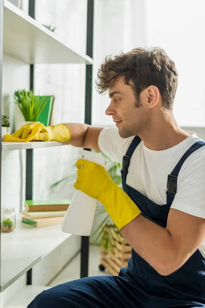 Vue latérale du bel homme en salopette nettoyage étagères rack dans l'appartement — Photo de stock