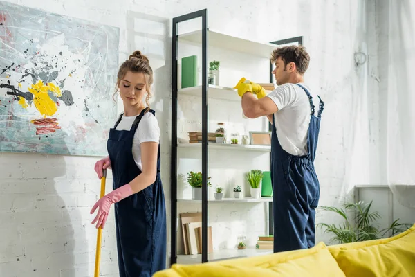 Attrayant femme en salopette nettoyage près beau nettoyant dans le salon — Photo de stock