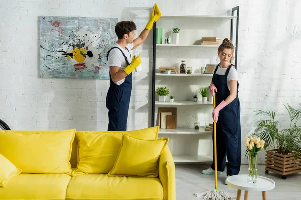 Attractive girl in overalls washing floor near handsome cleaner in living room — Stock Photo