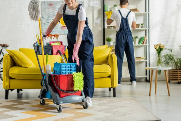 Vue recadrée de fille heureuse en salopette debout près du chariot de nettoyage — Photo de stock