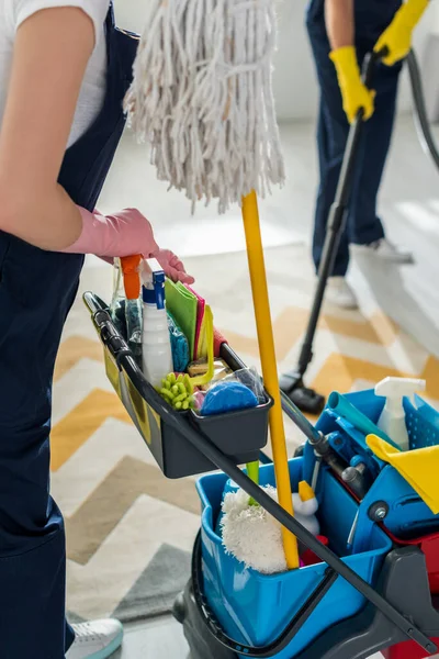 Vue recadrée du nettoyeur dans des gants en caoutchouc debout près du chariot de nettoyage et collègue avec aspirateur — Photo de stock