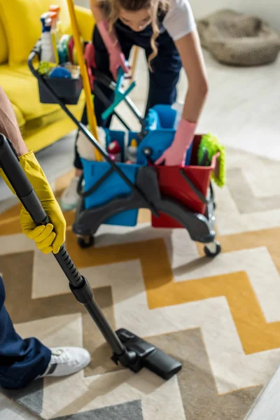 Selective focus of cleaner in rubber gloves standing near cleaning trolley and coworker with vacuum cleaner — Stock Photo