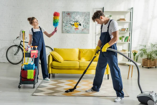 Limpiadores jóvenes en overoles haciendo limpieza de primavera de la sala de estar moderna - foto de stock