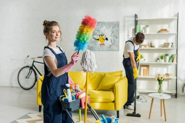 Enfoque selectivo de limpiador joven y feliz sosteniendo cepillo de plumero cerca del carro de limpieza — Stock Photo