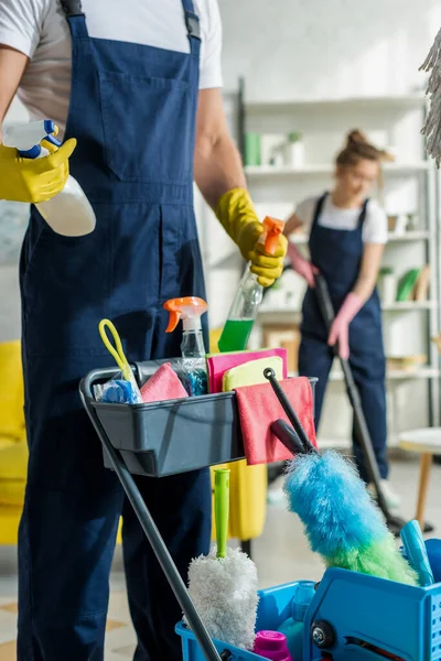 Selective focus of cleaner in uniform holding spray bottle near cleaning trolley — Stock Photo