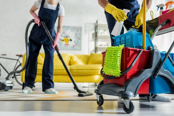 Vista ritagliata di detergente in guanti di gomma in possesso di bottiglia spray vicino al carrello di pulizia e collega con aspirapolvere — Foto stock