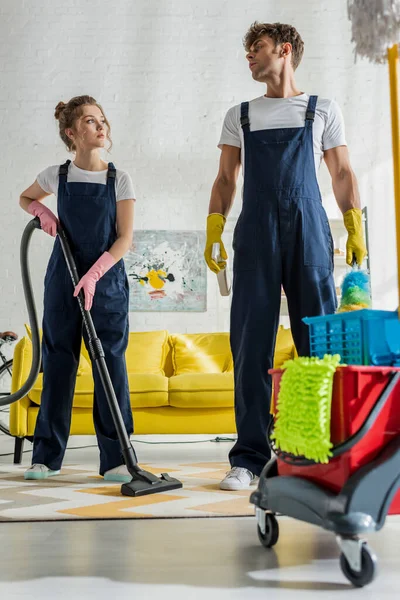 Selective focus of cleaners in uniform standing near cleaning trolley in modern living room — Stock Photo