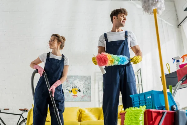 Foco seletivo de produtos de limpeza felizes em uniforme olhando para longe perto do carrinho de limpeza na sala de estar moderna — Fotografia de Stock