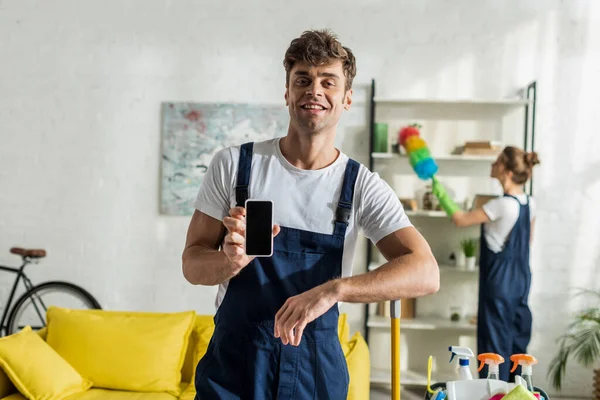 Enfoque selectivo de limpiador feliz y guapo celebración de teléfono inteligente con pantalla en blanco - foto de stock