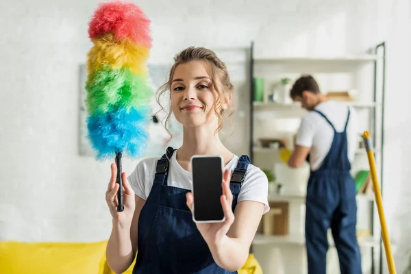 Selective focus of happy and attractive cleaner holding smartphone with blank screen and duster brush — Stock Photo