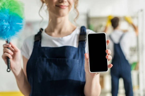 Mise au point sélective de la femme heureuse en uniforme tenant smartphone avec écran vierge et brosse à poussière — Photo de stock