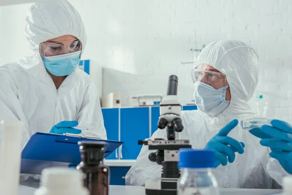 Foco seletivo de dois bioquímicos segurando placas de Petri perto do microscópio e recipientes médicos — Fotografia de Stock
