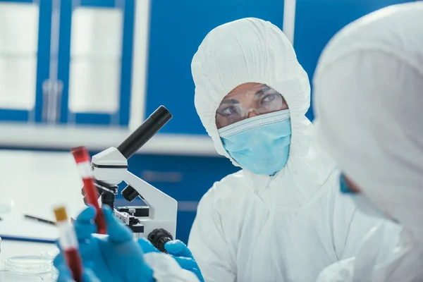 Biochemist in hazmat suit looking at colleague holding test tubes with blood samples — Stock Photo