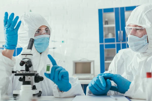 Dois bioquímicos em ternos hazmat falando em laboratório perto do microscópio — Fotografia de Stock