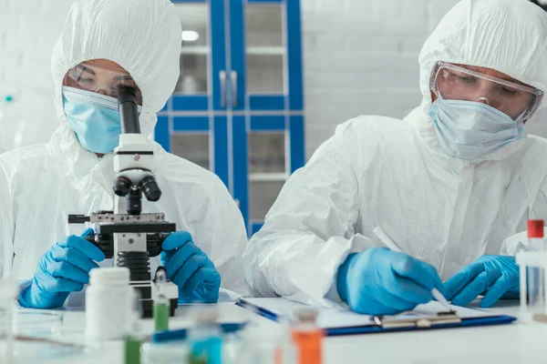 Selective focus of biochemist looking through microscope near colleague writing down results on clipboard — Stock Photo