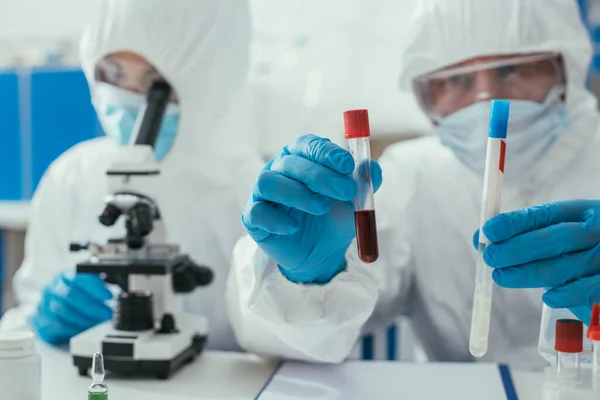 Selektiver Fokus des Biochemikers, der Reagenzgläser mit Flüssigkeits- und Blutprobe in der Nähe eines Kollegen hält, der durch das Mikroskop schaut — Stockfoto