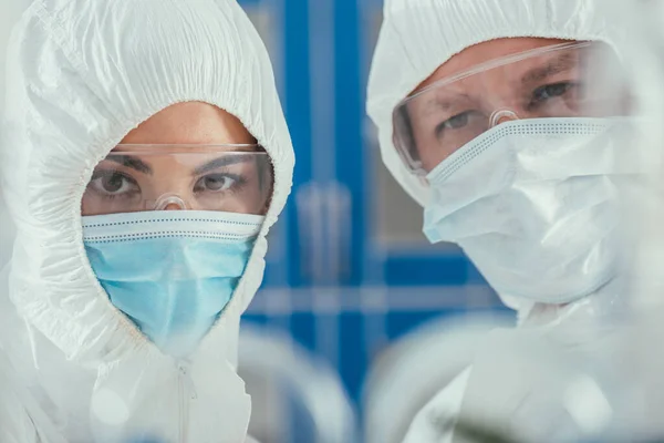 Enfoque selectivo de bioquímicos en suites de materiales peligrosos, máscaras médicas y gafas mirando a la cámara - foto de stock