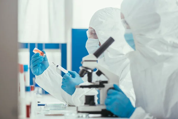 Foyer sélectif om biochimiste regarder à travers le microscope près collègue prenant des médicaments avec seringue — Photo de stock